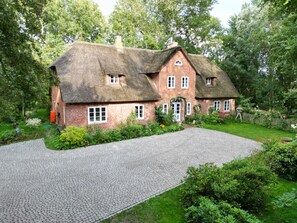 Ein traditionelles Backsteinhaus mit Strohdach, umgeben von üppigem Grün und einer gepflasterten Auffahrt.