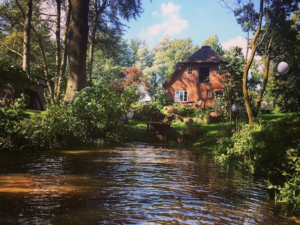 Blick auf ein traditionelles Backsteinhäuschen mit Strohdach, umgeben von üppigem Grün und Bäumen.