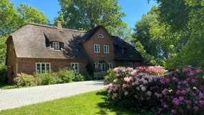 Ein Backsteinhaus mit Strohdach, einem gepflasterten Einfahrt und einem gepflegten Garten mit rosa Blumen.