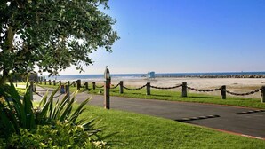 A paved pathway leading to a beach with a chain-link fence and a lifeguard tower in the distance.