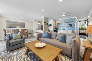 A modern living room with a grey sofa, a wooden coffee table, and a kitchen area with white cabinets and blue backsplash.