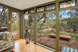 A room with a wooden floor, a floral sofa, a small table with a plant, and large windows offering a view of a garden and mountains. - ☼Views! Private House, Hot Tub, Hiking, Fireplace, Art☼ (Sedona)