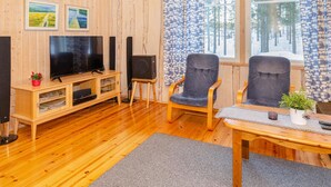 A cozy living room with wooden walls, a TV, two chairs, a small table, and a window with curtains.