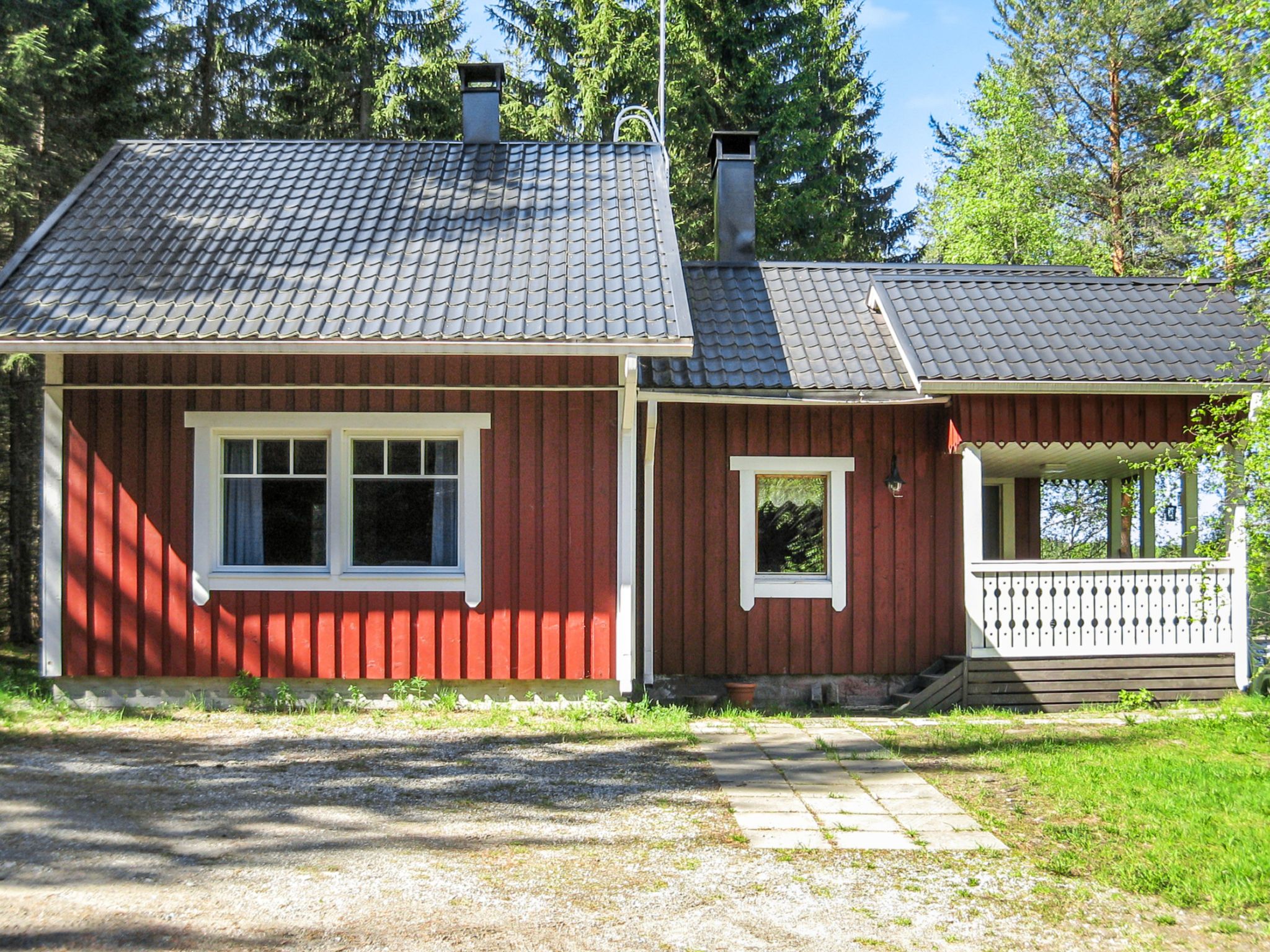 A red wooden cabin with a grey tiled roof, a small porch, and a gravel driveway.