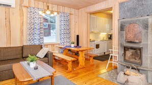 A cozy cabin interior with a wooden floor, a sofa, a dining table with benches, a kitchen area, and a stone fireplace.