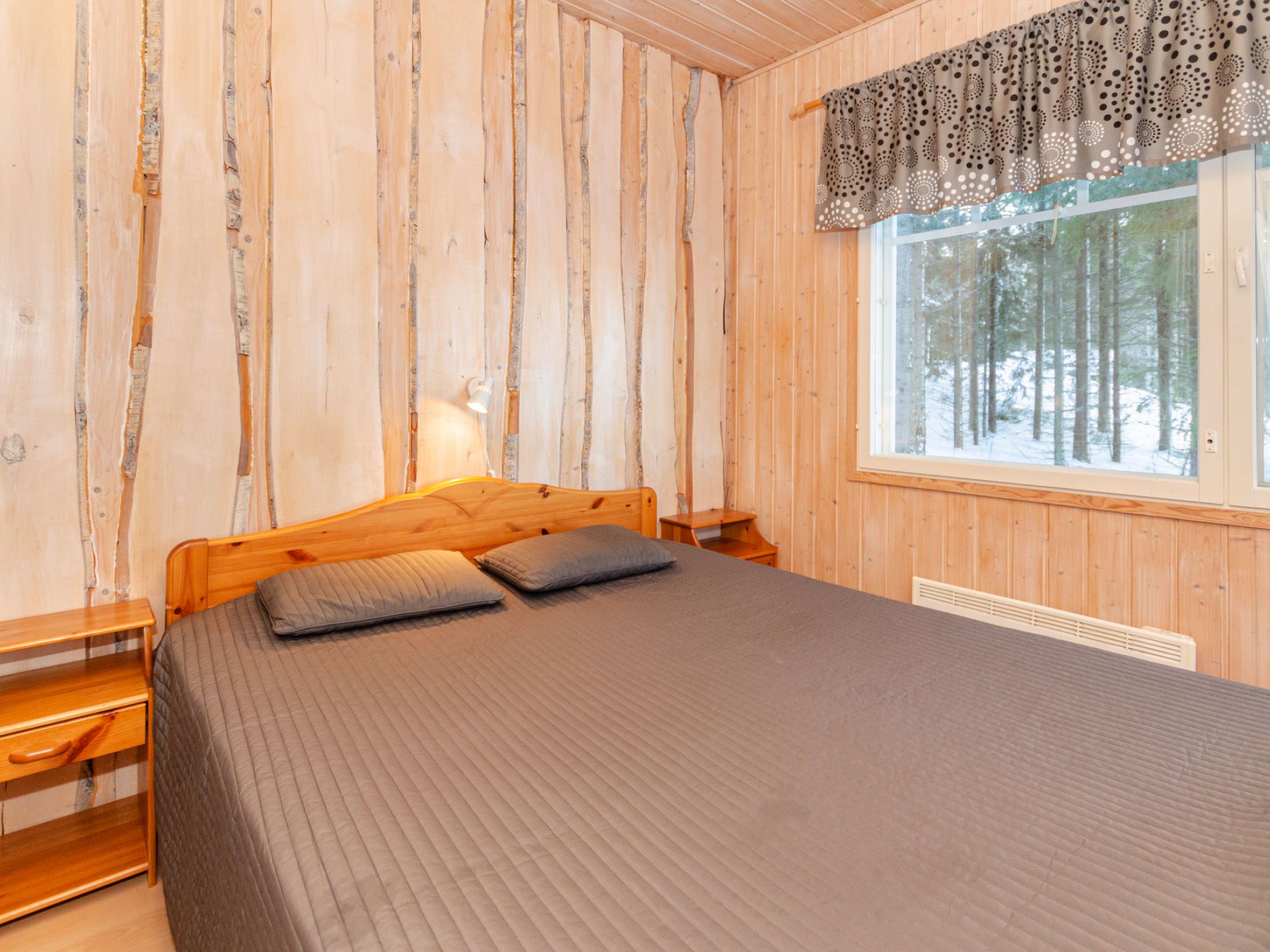 Ein Schlafzimmer einer Holzhütte mit einem Bett, Kissen, einem Nachttisch, einer Lampe und einem Fenster mit Blick auf einen verschneiten Wald.