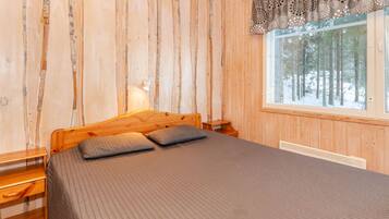 Ein Schlafzimmer einer Holzhütte mit einem Bett, Kissen, einem Nachttisch, einer Lampe und einem Fenster mit Blick auf einen verschneiten Wald.