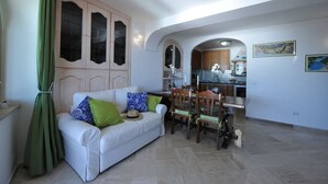 A living room with a white sofa, green and blue pillows, a dining table with chairs, and a kitchen in the background. - Casa Carola beautiful sea view and relax !!!! (Positano)