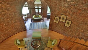 Um amplo loft com pé direito alto, paredes de tijolo aparente, área de estar central com sofá e banquinho, cadeira balanço e escada para um nível superior.