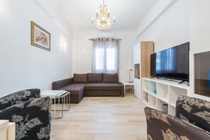 Un salon moderne avec un canapé d’angle, un téléviseur à écran plat, un lustre et un tableau accroché au mur.