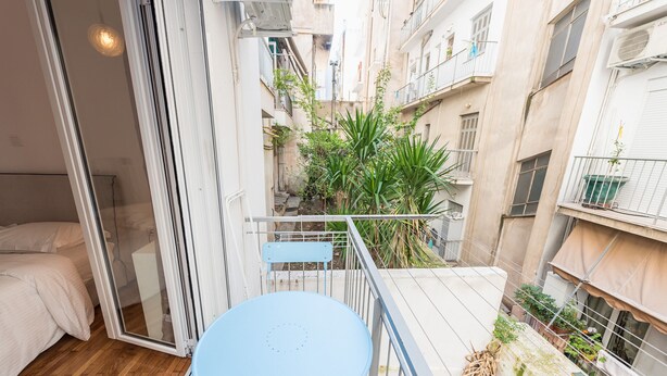 A small balcony with a blue table and chair, a bed, and a view of a narrow alley with plants and buildings. - Fresh and Clean apt for 6 in Koukaki (Athens)