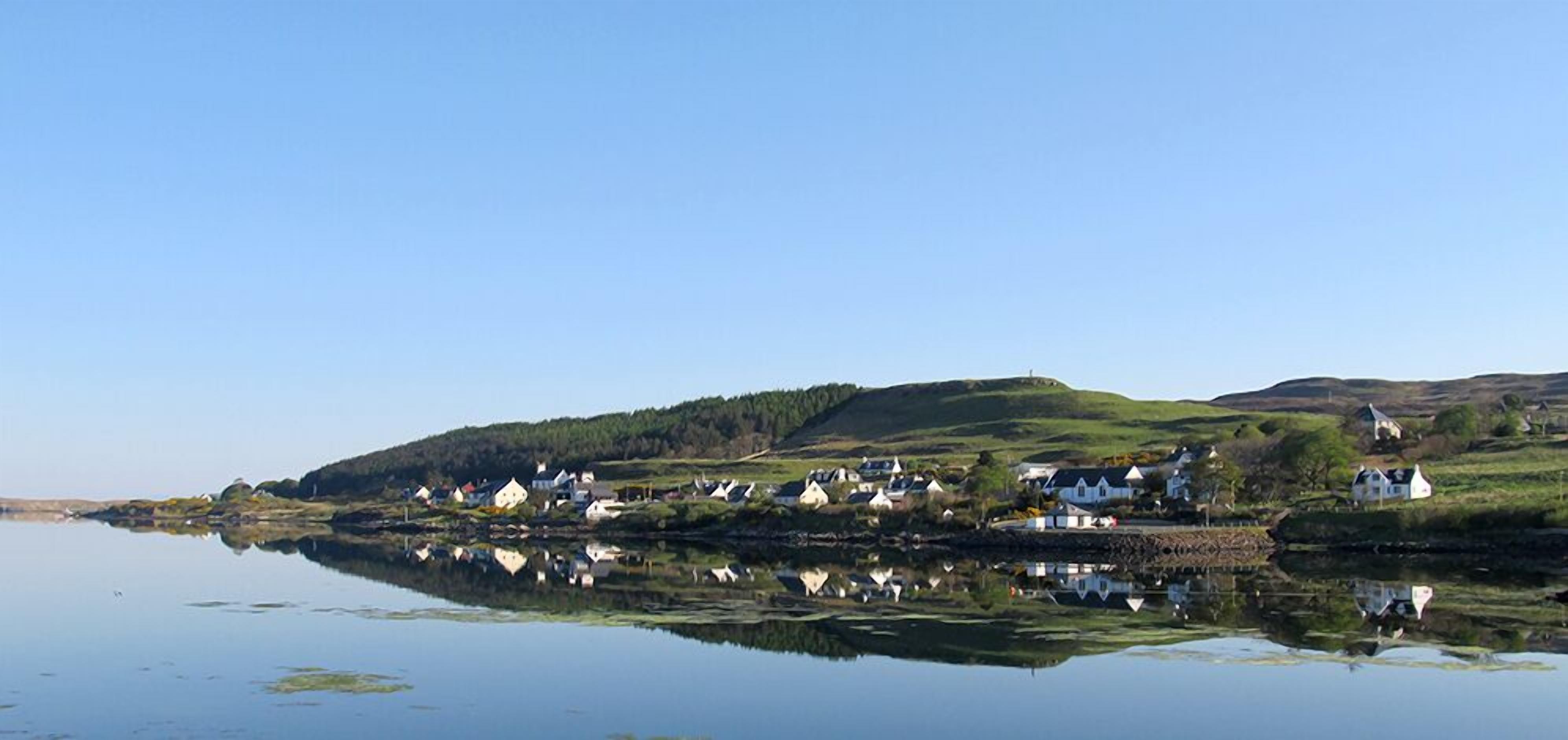Un village côtier avec des maisons qui bordent le rivage, un lac paisible dans lequel se reflètent les bâtiments, et une colline en arrière-plan.