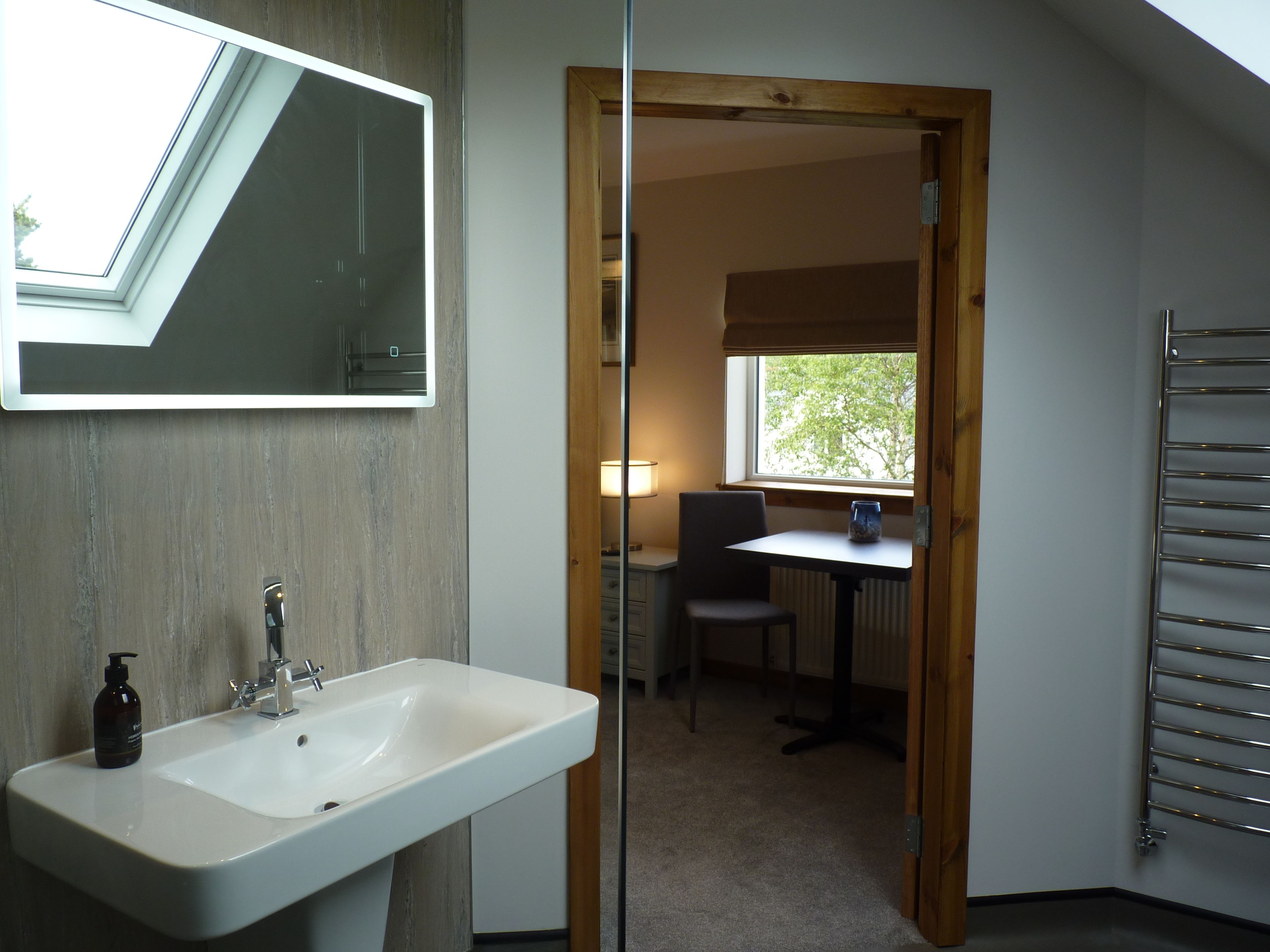 Une salle de bain avec un lavabo blanc, un miroir et un flacon de savon. Il y a une fenêtre donnant sur des arbres et une porte qui mène à une pièce où se trouvent une table et une chaise.