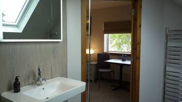 Une salle de bain avec un lavabo blanc, un miroir et un flacon de savon. Il y a une fenêtre donnant sur des arbres et une porte qui mène à une pièce où se trouvent une table et une chaise.
