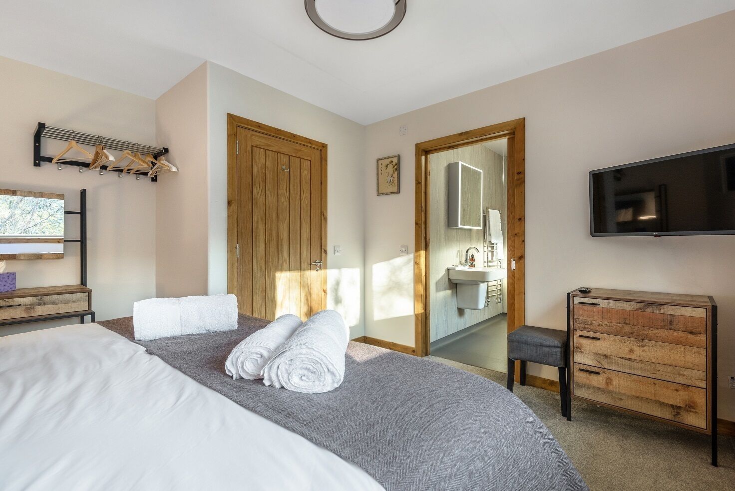 A bedroom with a bed, a wooden dresser, a TV, a mirror, and a bathroom visible through an open door.