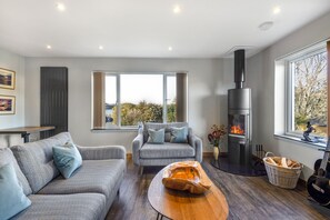 Ein modernes Wohnzimmer mit Kamin, grauen Sofas, einem Couchtisch und Blick durch große Fenster.