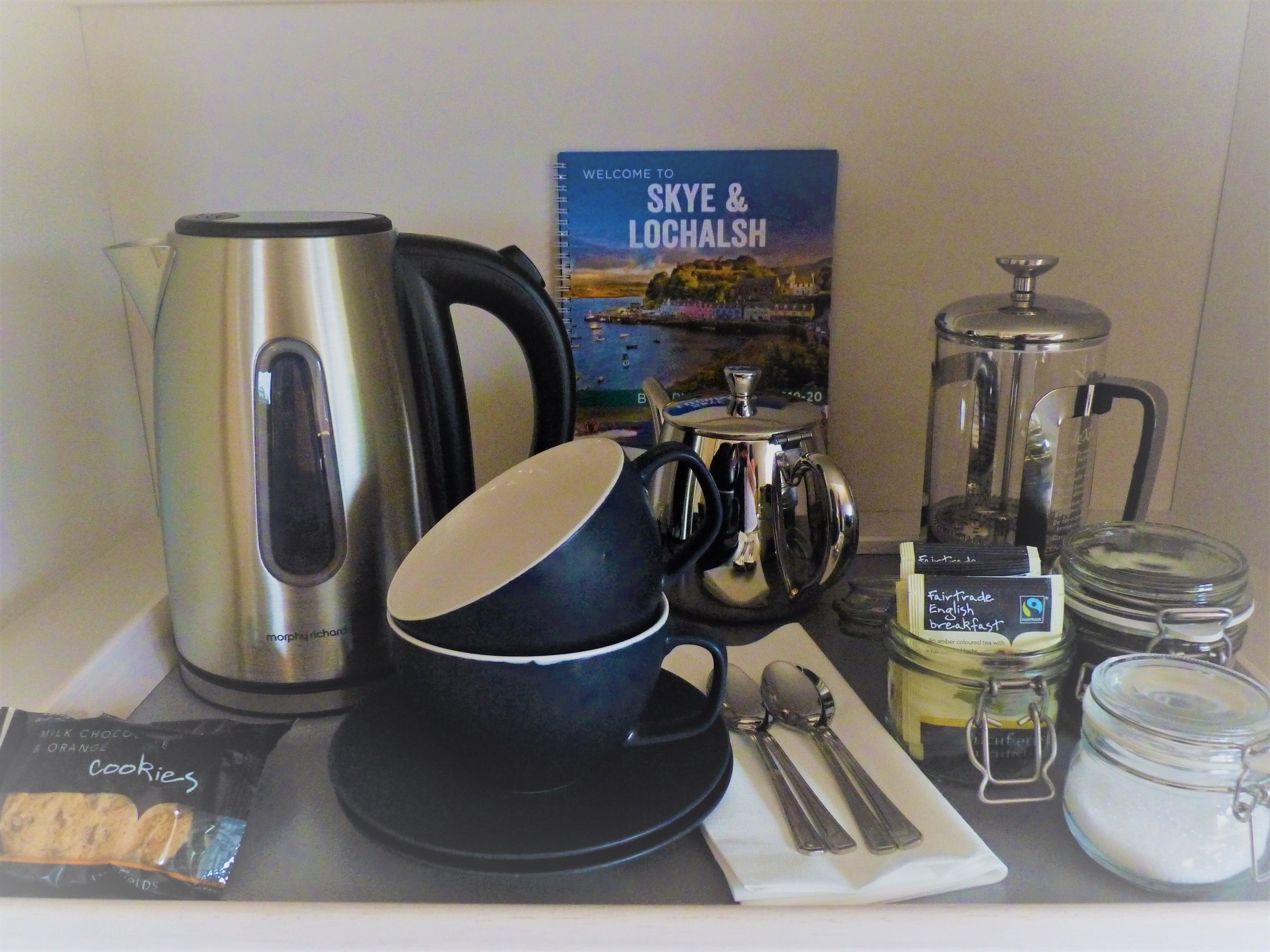 A kitchen counter with a kettle, a coffee pot, a book titled "Welcome to Skye & Lochalsh", a jar of sugar, and a packet of cookies.