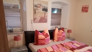 Ein Schlafzimmer mit einem Bett, zwei Nachttischen, einem Fenster mit Jalousien und einem an der Wand hängenden Kunstwerk.