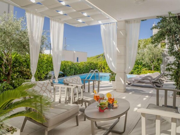 A covered outdoor seating area with a pool, lounge chairs, and a table with drinks and fruit.