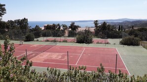 A tennis court with a net, surrounded by trees and a view of the sea and hills.