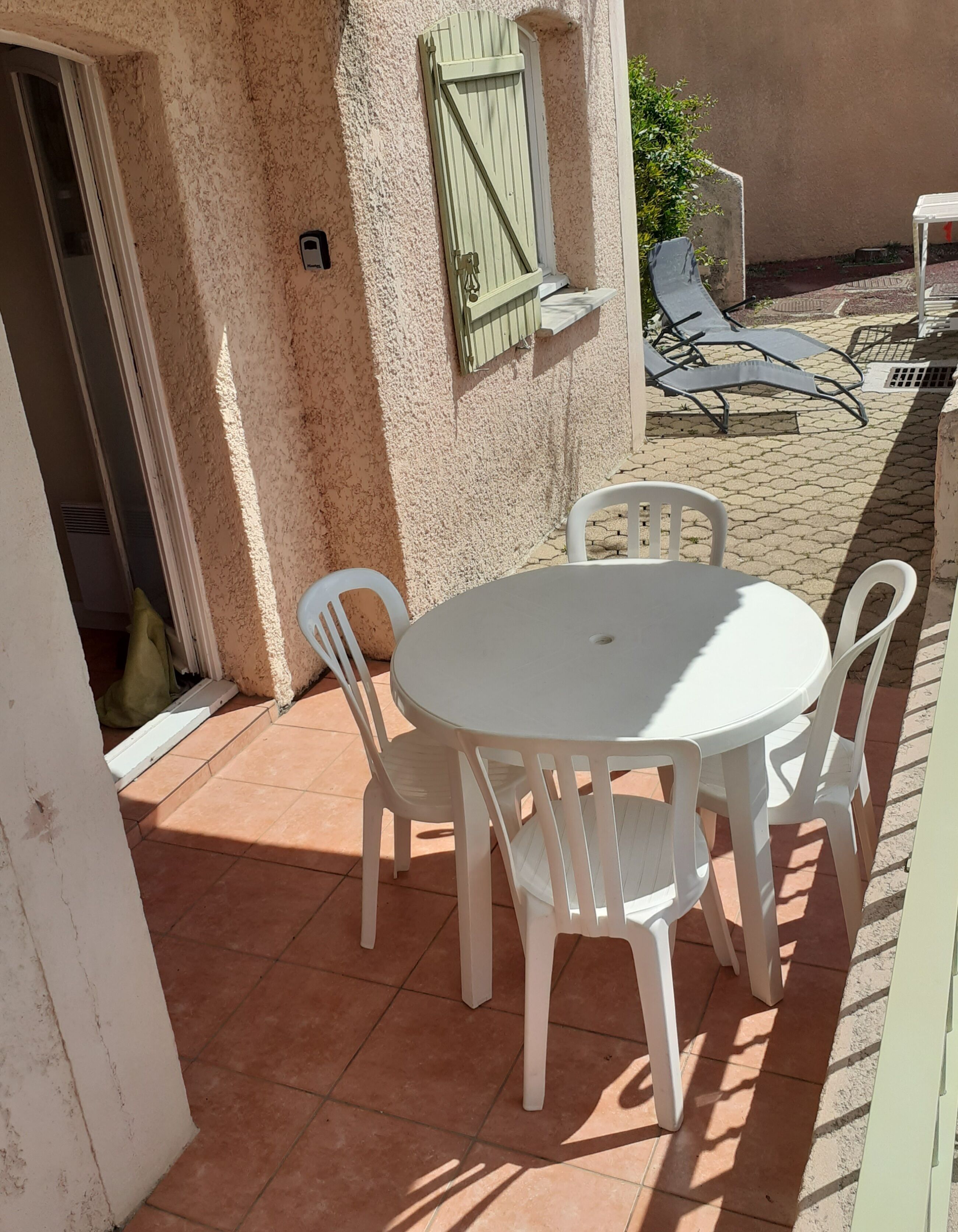 Een terras met een ronde tafel en vier stoelen, een deur en een raam met een luik.