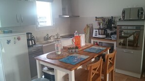 Een compacte keuken met een koelkast, fornuis, magnetron en gootsteen. Boven de gootsteen bevindt zich een raam en er staat een houten tafel met stoelen.