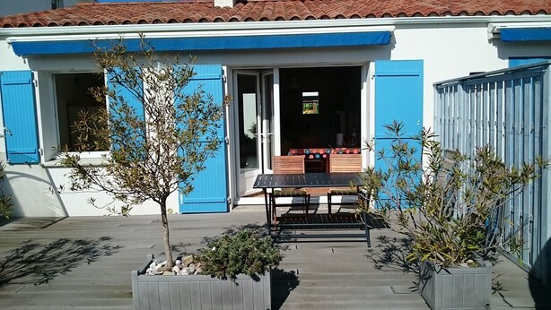 Een huis met blauwe luiken, een terras met een tafel en stoelen, en potplanten.