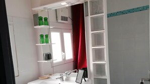Un bagno con un lavandino bianco, uno specchio e mensole con bottiglie verdi. Si vede una tenda rossa, un cestino dei rifiuti blu e una doccia con piastrelle blu.