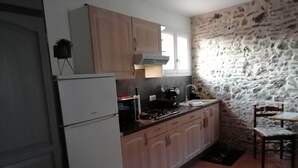 Una cucina compatta con frigorifero, fornello e lavello. Presenta mobili in legno chiaro, una parete in pietra e un piccolo angolo ristoro con una sedia.