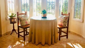 Una mesa redonda con mantel blanco, dos sillas de madera con cojines estampados y una planta en maceta sobre una mesita junto a la ventana.