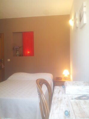 Quarto de hotel com cama, uma cadeira de madeira, mesa de cabeceira com abajur e cortina vermelha na parede.