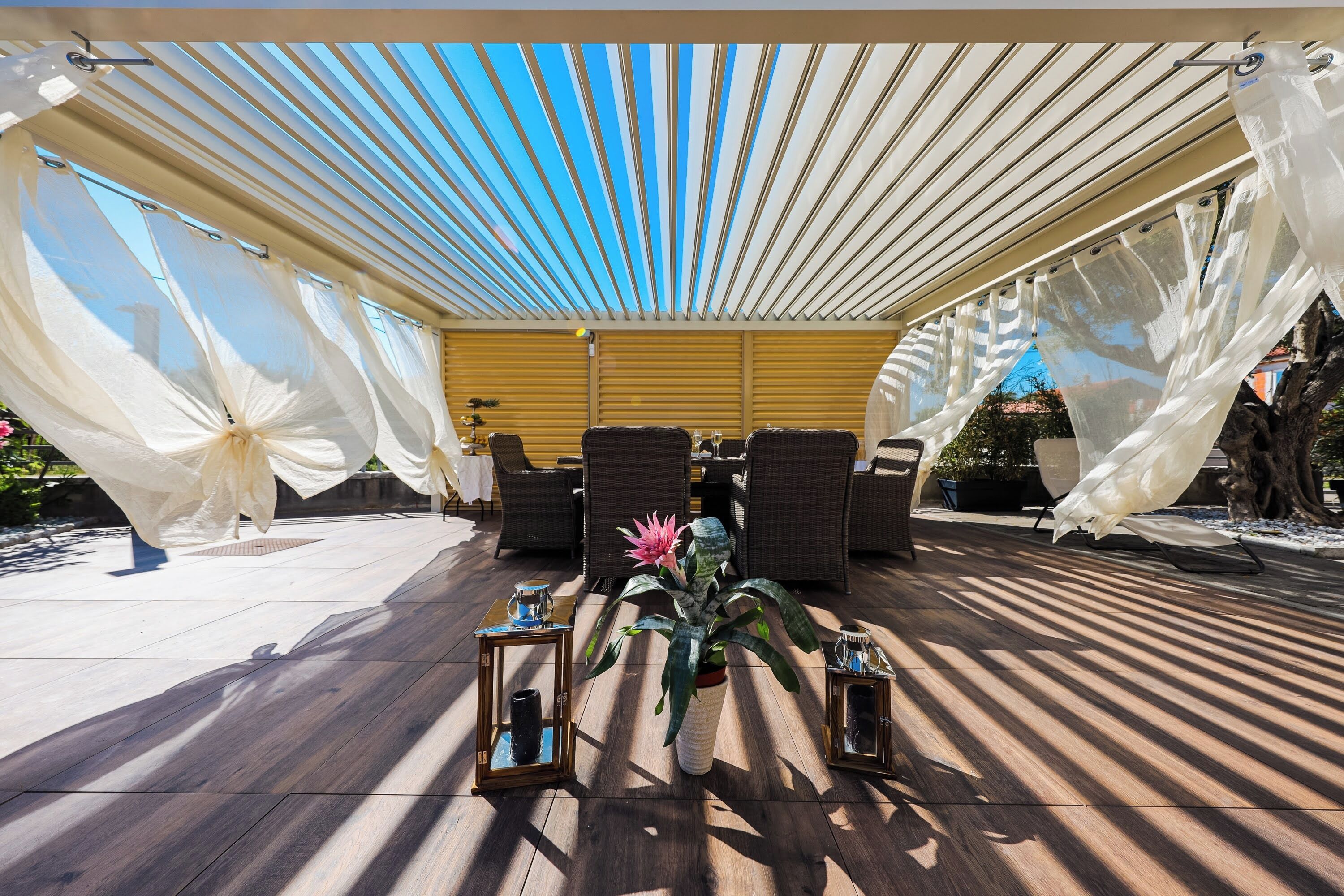 Eine Terrasse im Freien mit Holzboden, weißen Vorhängen, Korbmöbeln und Topfpflanzen.