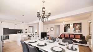 Eine moderne Küche mit weißen Schränken, ein Essbereich mit einem weißen Tisch, gedeckt für sechs Personen, und eine Wohnbereich mit einem schwarzen Sofa und roten Kissen.