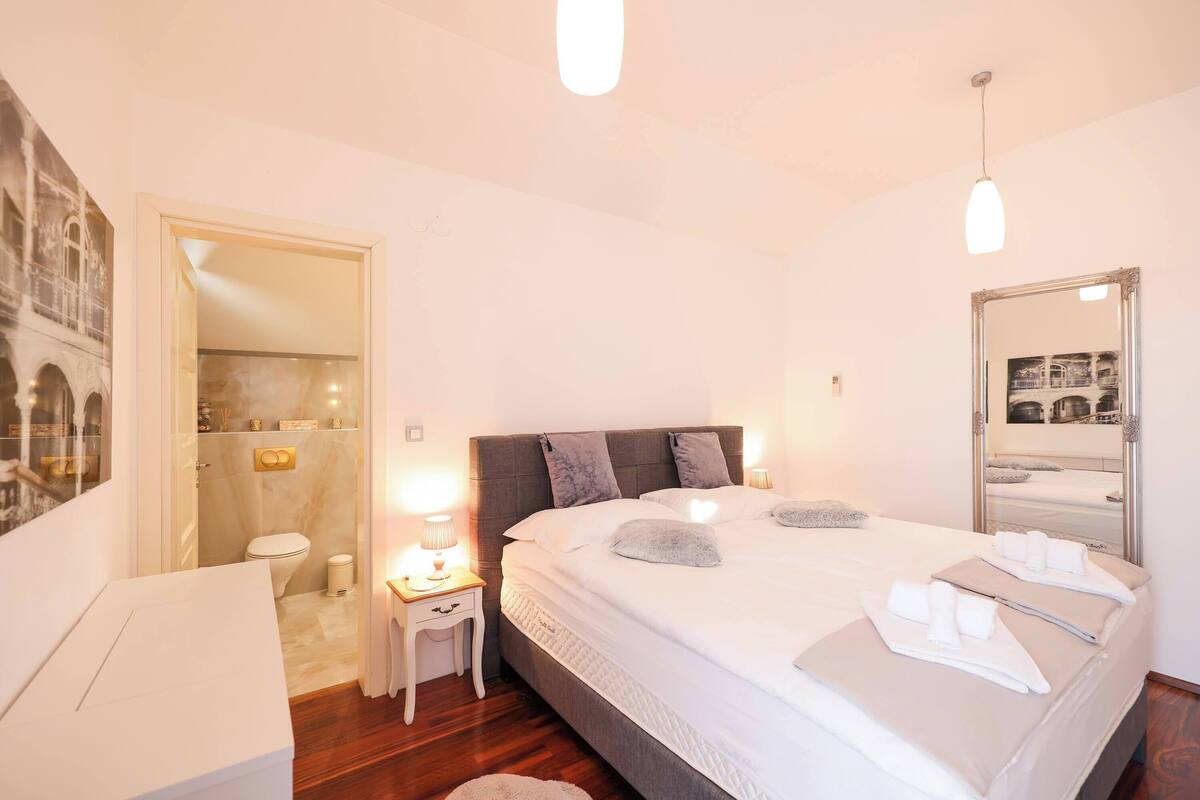 A neatly arranged bedroom with a large bed, bedside tables, a mirror, and a bathroom visible through an open door.