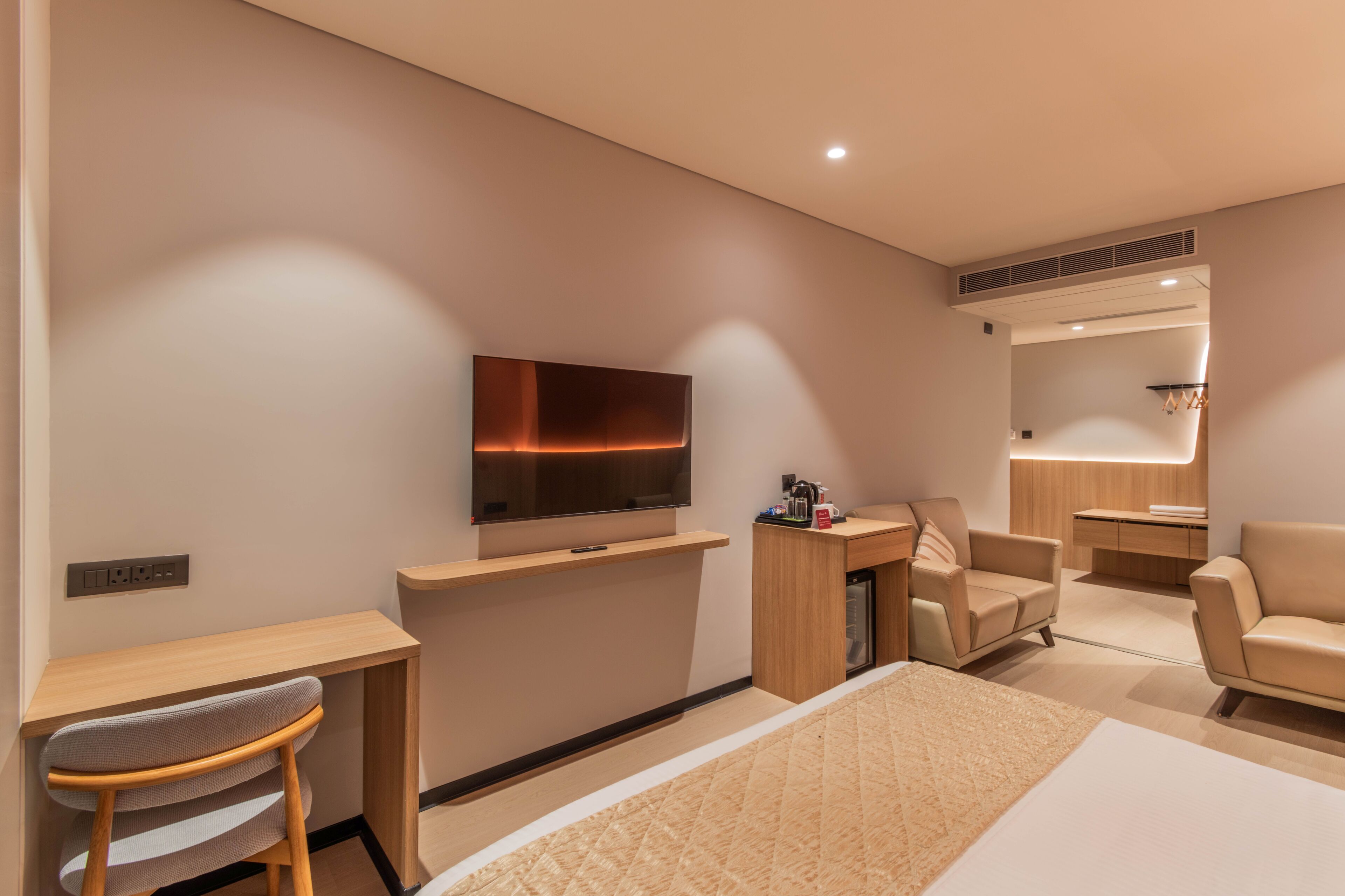 A modern hotel room with a bed, a desk, a chair, a television, and a small kitchenette area.