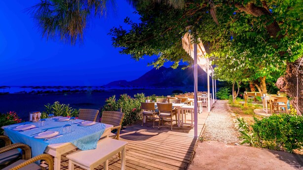 An outdoor dining area with tables and chairs, overlooking a body of water and mountains.