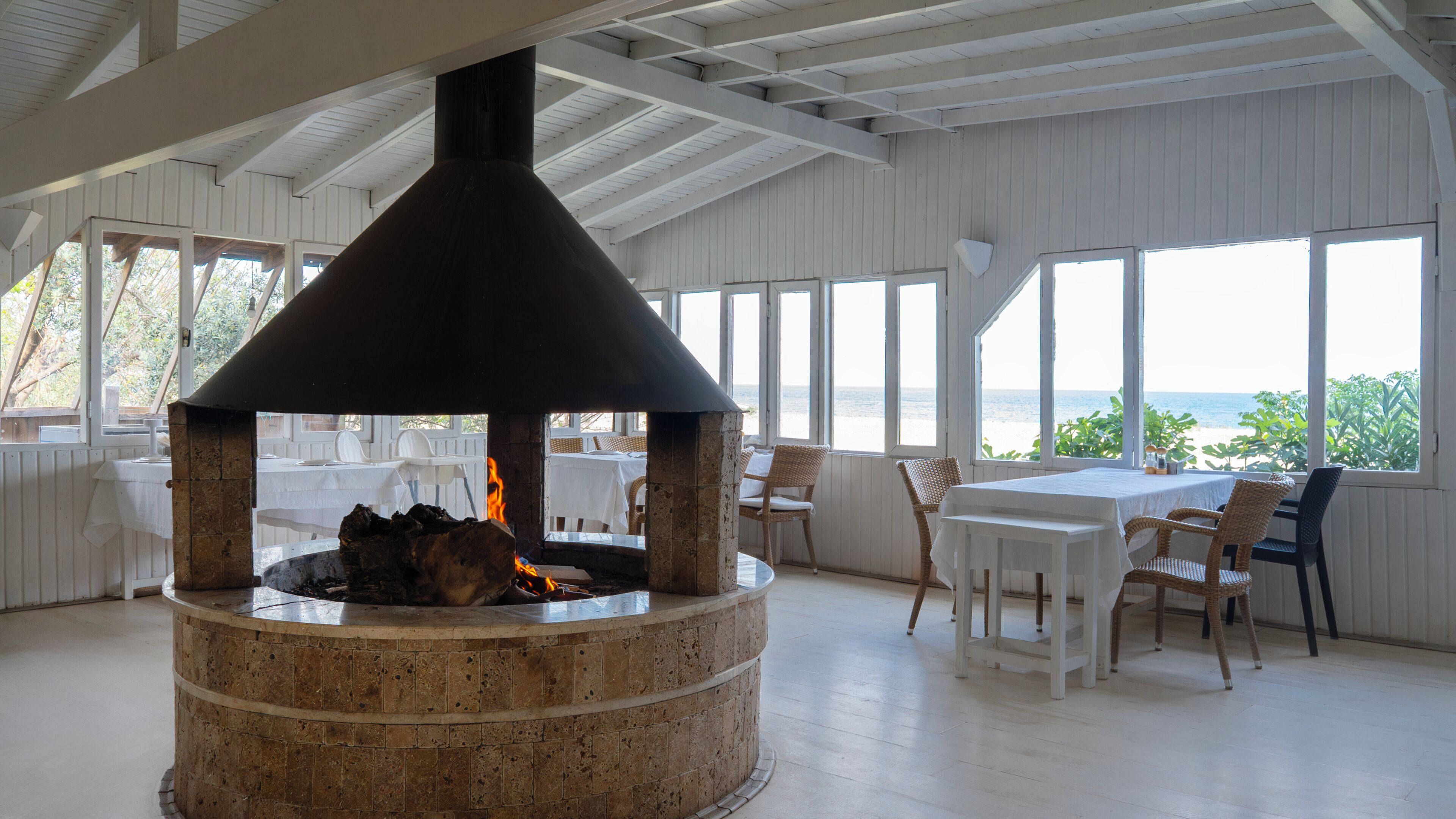 Un foyer en pierre avec une cheminée, entouré de chaises en osier blanc et d’une table, avec vue sur la mer à travers de grandes fenêtres.