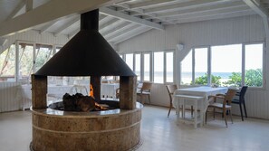 A stone fire pit with a chimney, surrounded by white wicker chairs and a table, with a view of the sea through large windows. - Etenna Beach Bungalows (Kemer)