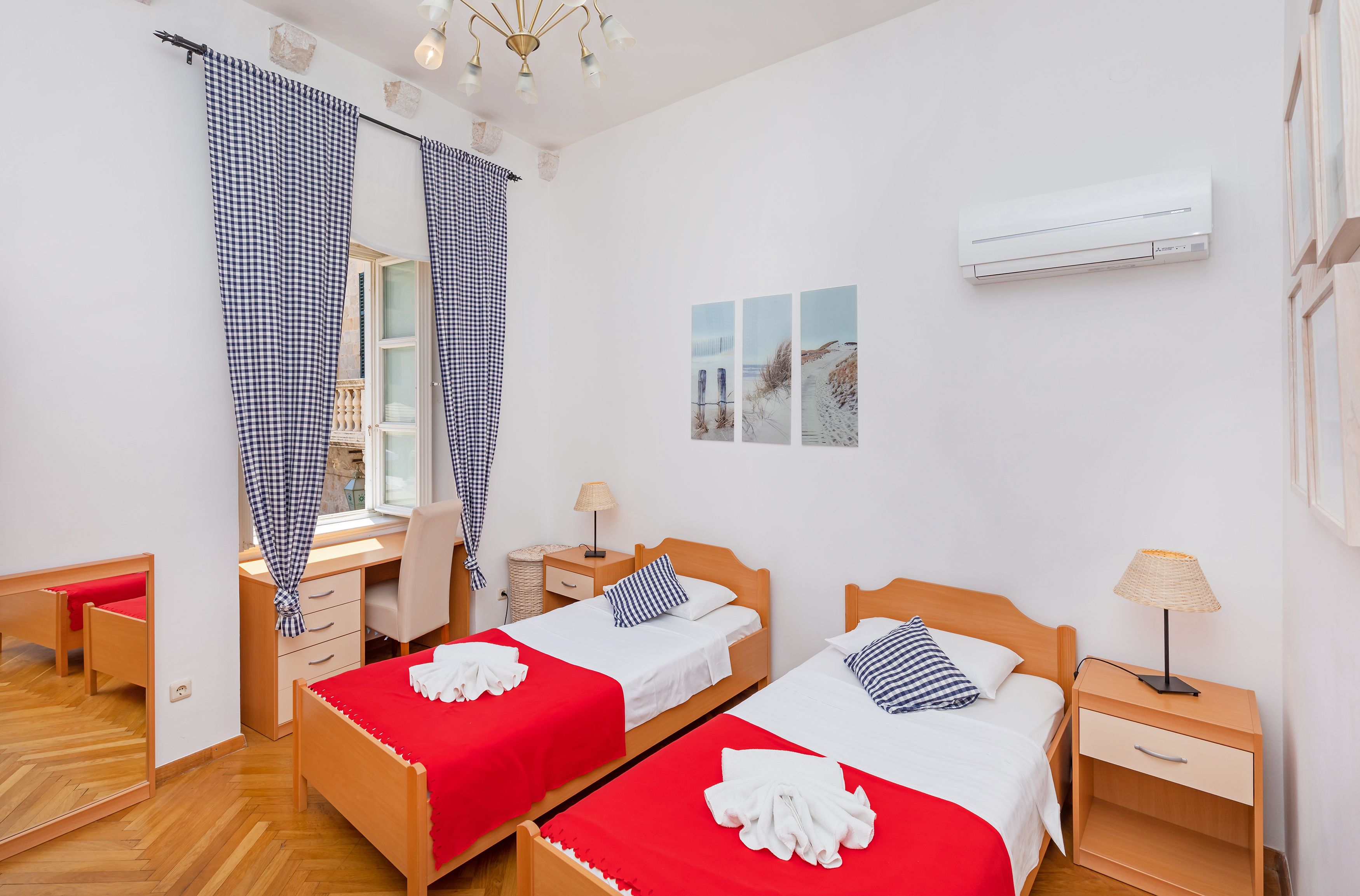 Habitación con dos camas, un escritorio y una silla. Se ve una ventana con cortinas, una lámpara y un aire acondicionado en la pared.
