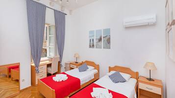 A room with two beds, a desk, and a chair. There is a window with curtains, a lamp, and an air conditioner on the wall.