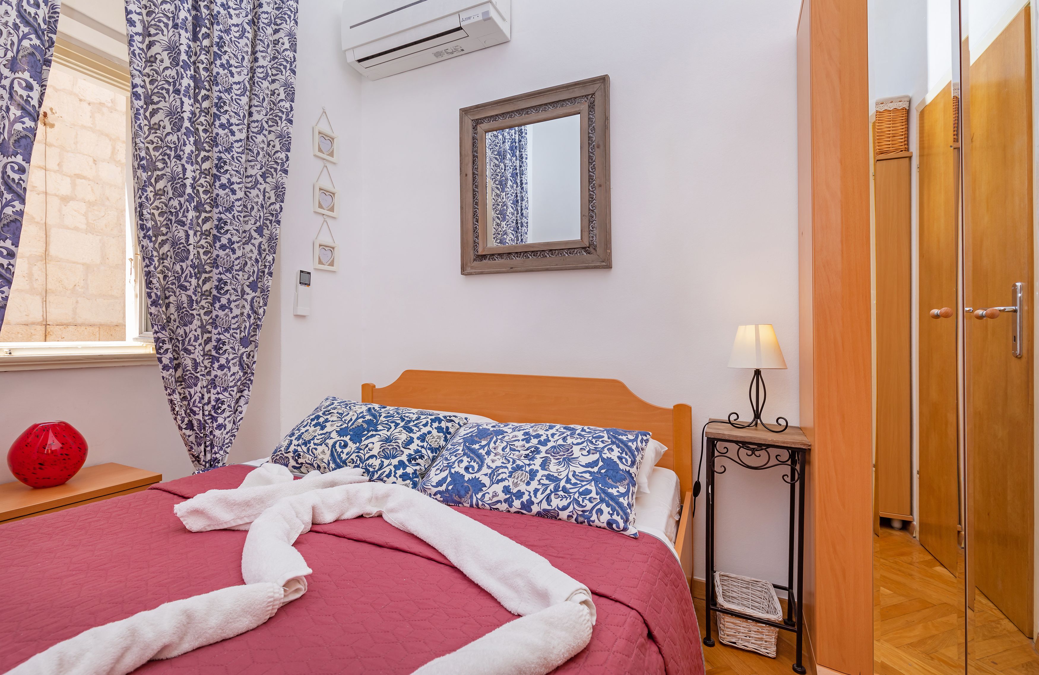 A bedroom with a bed, a bedside table with a lamp, a wall-mounted air conditioner, and a wooden wardrobe.