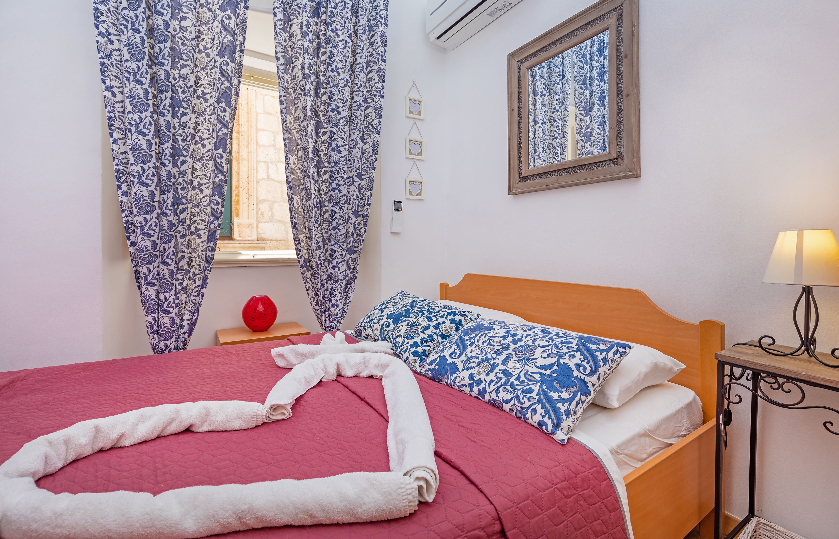 Een keurig opgemaakt bed met een rode sprei, kussens met een blauw-witte print, en een witte handdoek gevouwen in de vorm van een hart.
