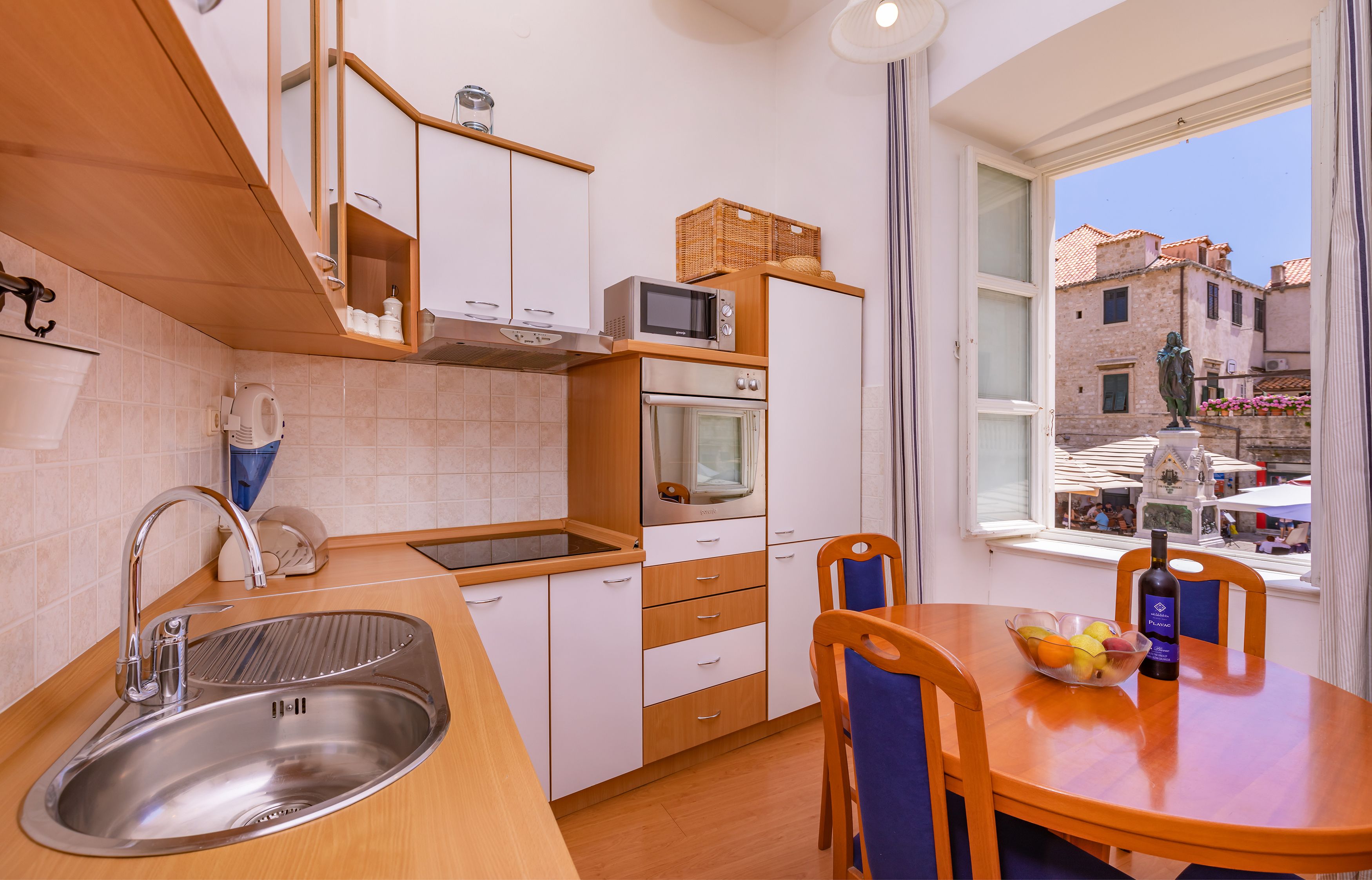 Una cocina moderna con un lavamanos de acero inoxidable, armarios de madera, un microondas y un horno. Hay una mesa de comedor con sillas, un bol de fruta y una botella de vino.