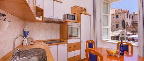 Una cocina moderna con un lavamanos de acero inoxidable, armarios de madera, un microondas y un horno. Hay una mesa de comedor con sillas, un bol de fruta y una botella de vino.
