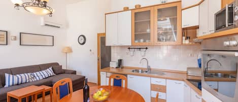 Una cocina compacta con armarios blancos, un microondas y un lavamanos. Hay una mesa de comedor con sillas y un bol de fruta.