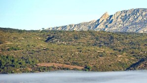 Paisagem montanhosa com um pequeno edifício em primeiro plano, rodeado por campos e folhagem outonal.