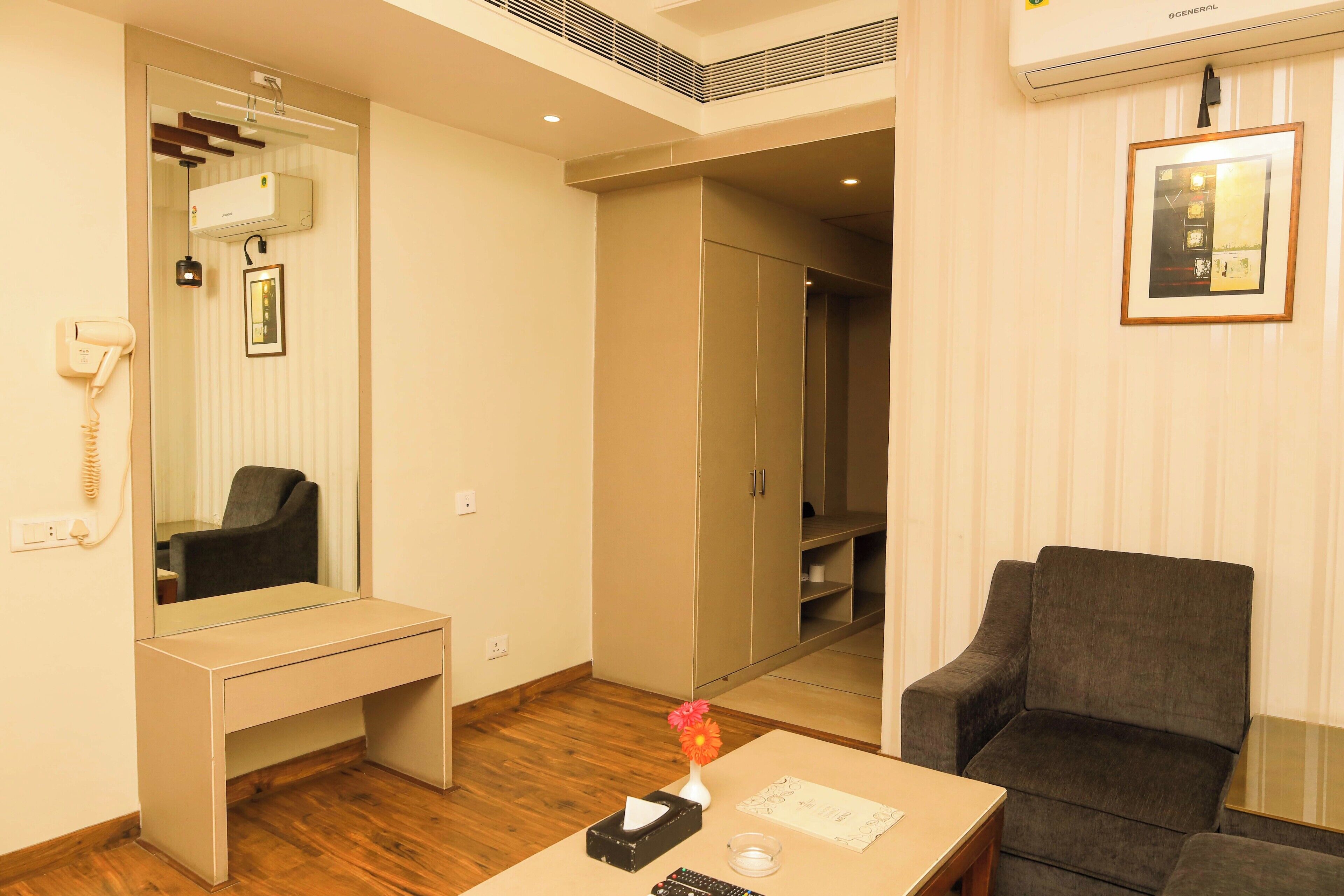 A hotel room with a desk, chair, mirror, wardrobe, and a small table with a vase and remote controls.