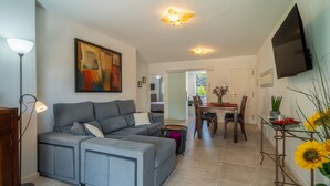 A modern living room with a grey sectional sofa, a dining area with a table and chairs, a flat-screen TV mounted on the wall, and a large painting on the wall.