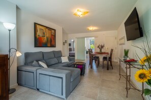 A modern living room with a grey sectional sofa, a dining area with a table and chairs, a flat-screen TV mounted on the wall, and a large painting on the wall.