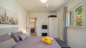 A bedroom with a bed, pillows, a ceiling fan, a window with curtains, a wall-mounted TV, and a door leading to another room.